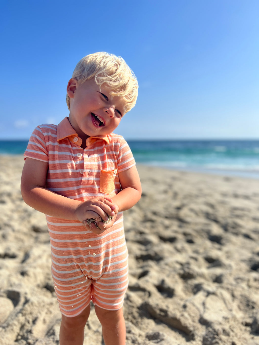 adorable blonde hair boy, wearing our peach/white polo romper , rolling a ball of sand in his hands at the beach in summer.