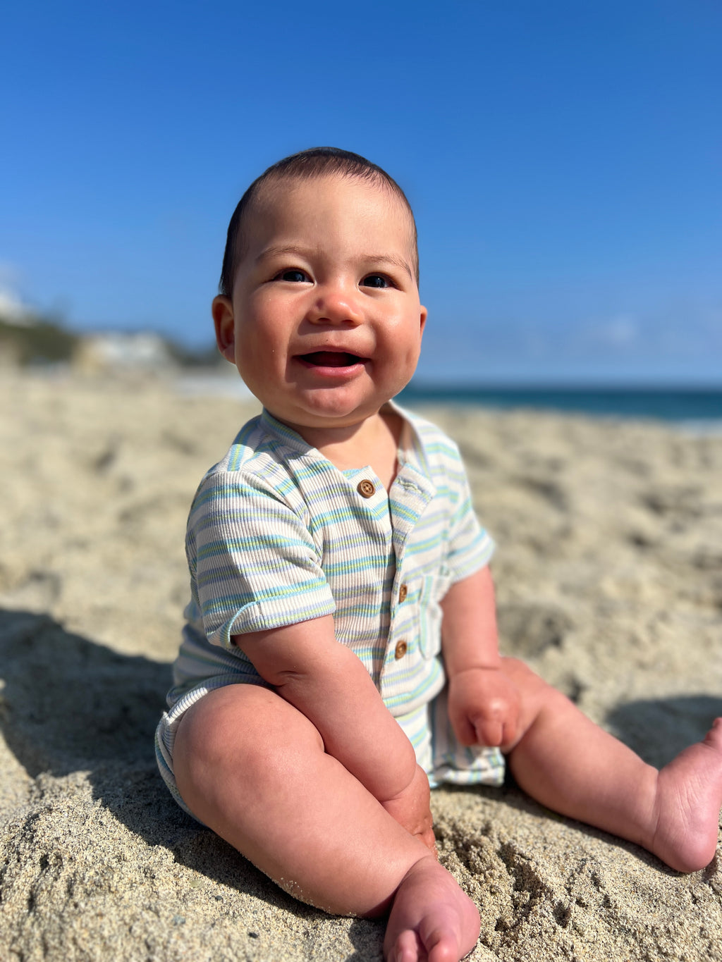 Adorable baby boy with brown hair and brown eyes, wearing our beige/lime/grey ribbed henley romper with a big smile, sitting on the sand at the beach in summer.