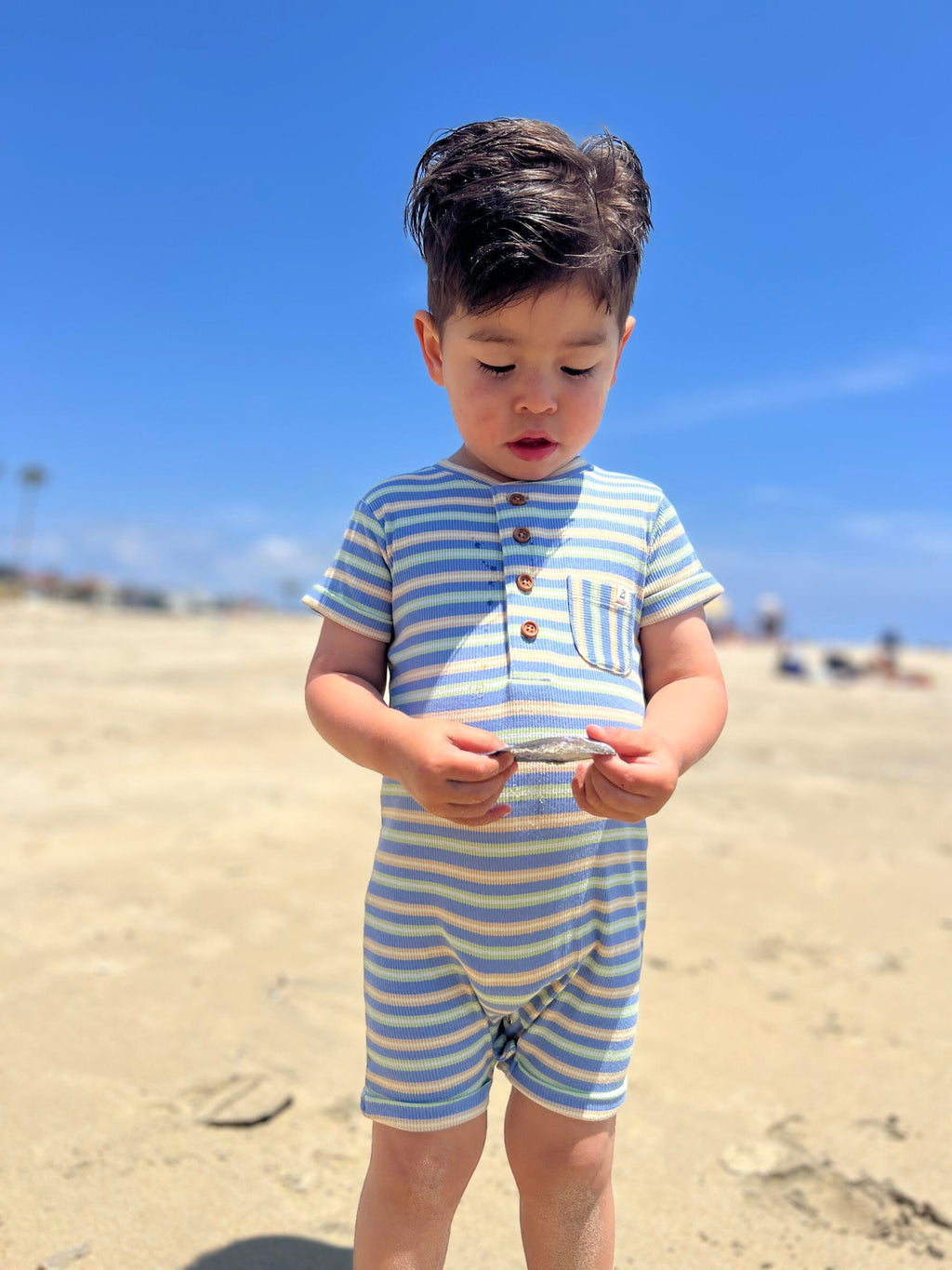 small boy with dark brown hair wearing our pink/white/grey ribbed henley romper standing on the beach holding a seashell found in the sand