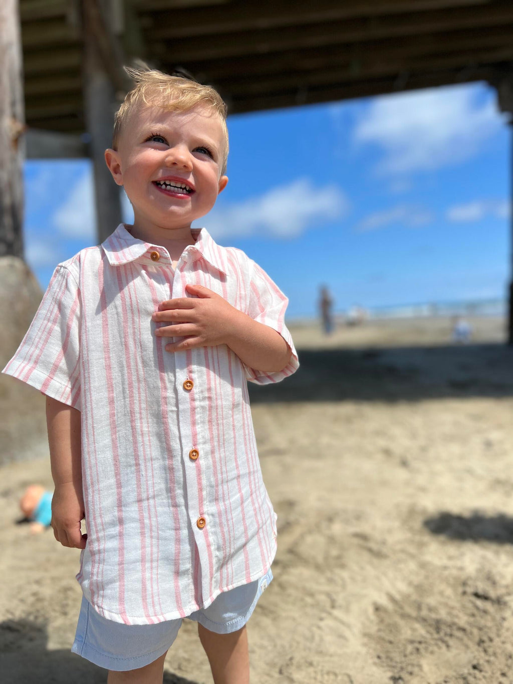 blonde hair boy with blue eyes wearing the pink/cream woven shirt and pale blue gauze shorts , standing at the beach under a wooden bridge.