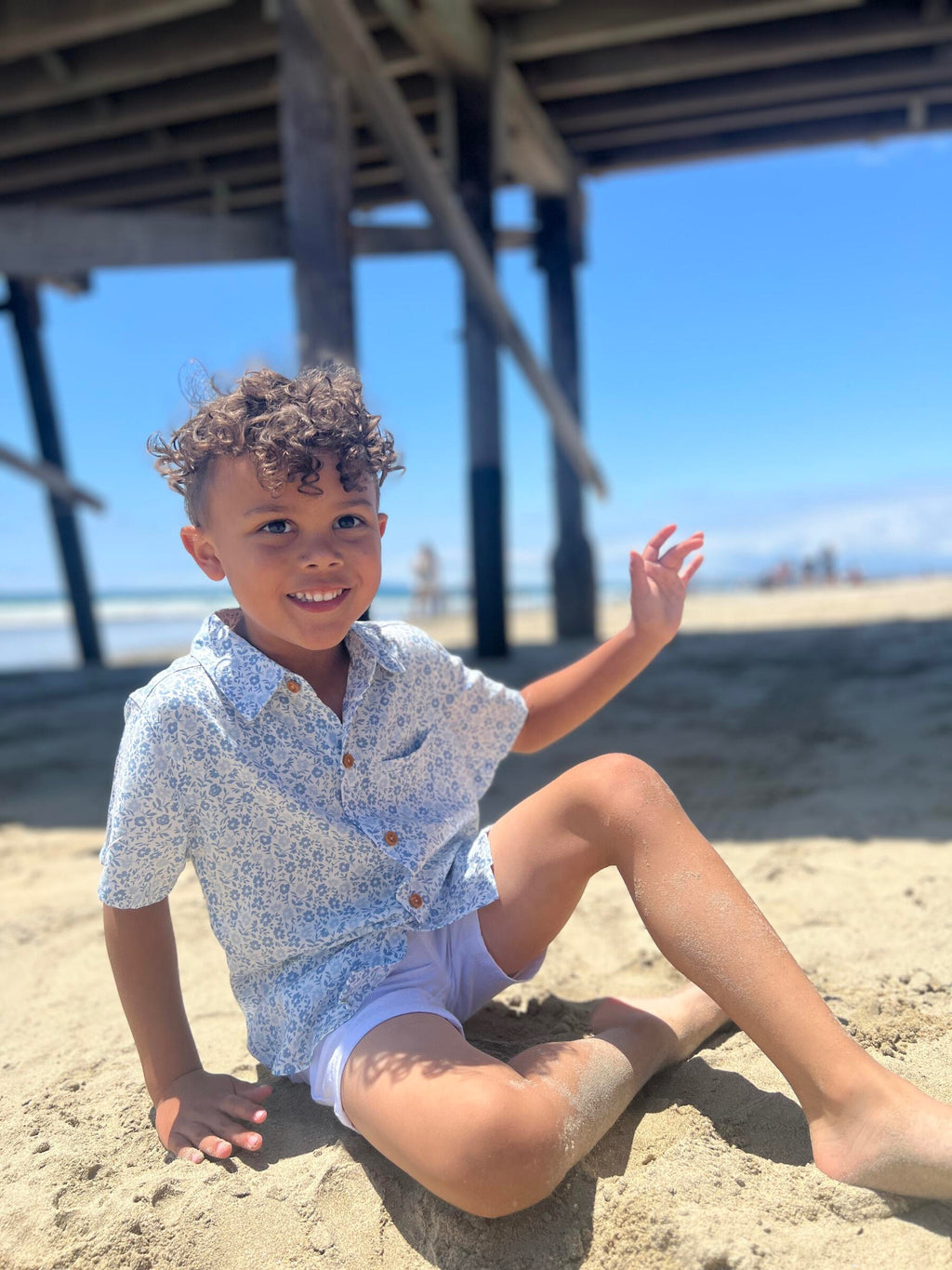 A boy with brown curly hair wearing the blue floral woven shirt and white gauze shorts, sitting on the sand at the beach.