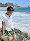 Boy with black hair, wearing our pale grey woven shirt with green palm woven shorts, walking across the small rocks pn the sea.
