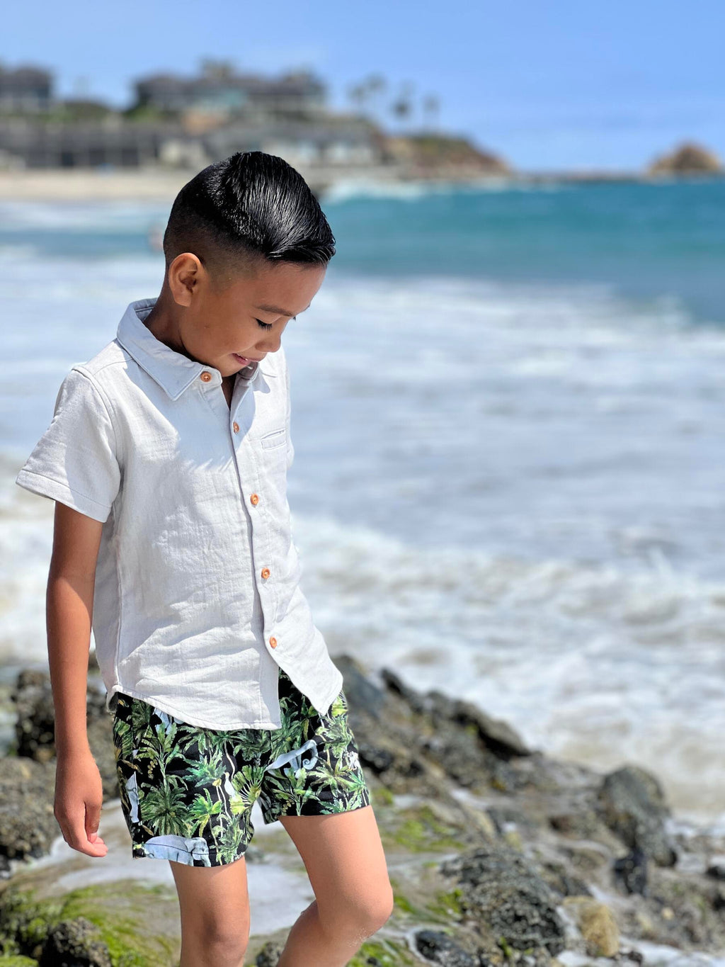 Boy with black hair, wearing our pale grey woven shirt with green palm woven shorts, walking across the small rocks pn the sea.