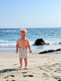 small blonde hair boy wearing the apricot woven shirt and cream shorts with removable suspenders walking by the sea.