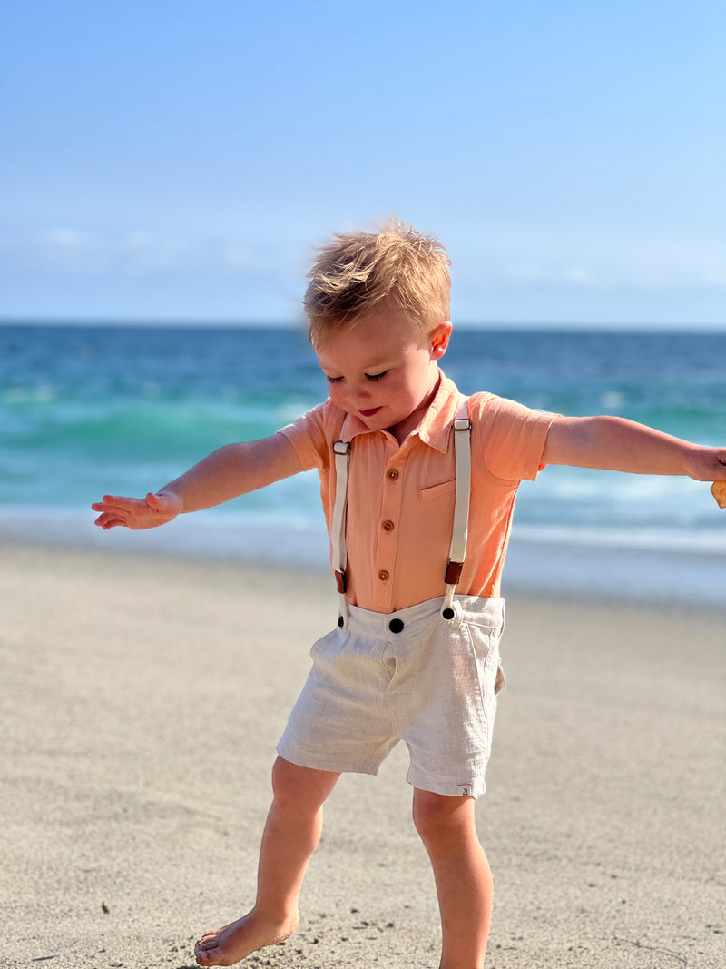 small blonde hair boy wearing the apricot woven shirt and cream shorts with removable suspenders walking by the sea.