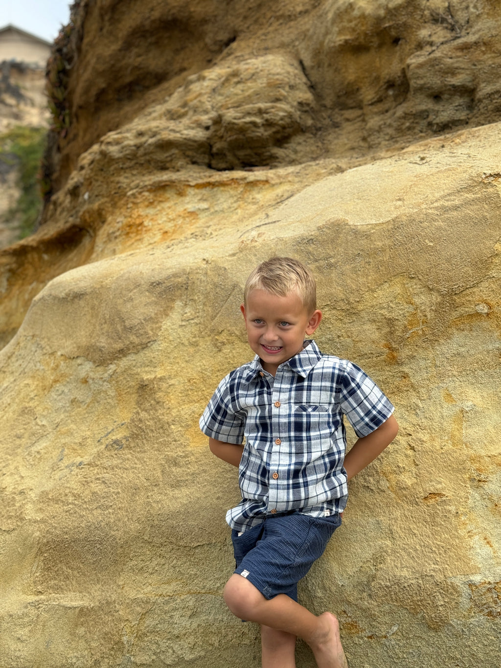 Blonde hair boy, with blue eyes wearing the navy/white plaid woven shirt and navy twill shorts posing in front of large beach rock.
