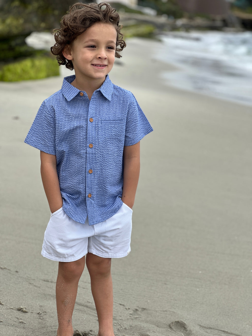Small boy with curly brown hair wearing our navy seersucker woven shirt and white gauze shorts with his hands in his pockets standing on the sand at the beach.