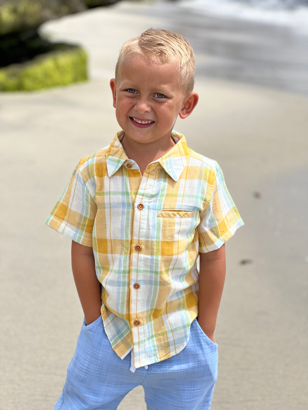 Blonde hair boy with blue eyes, wearing the gold/cream plaid woven shirt and blue heather pants, with his hands in the pocket, smiling at the camera.
