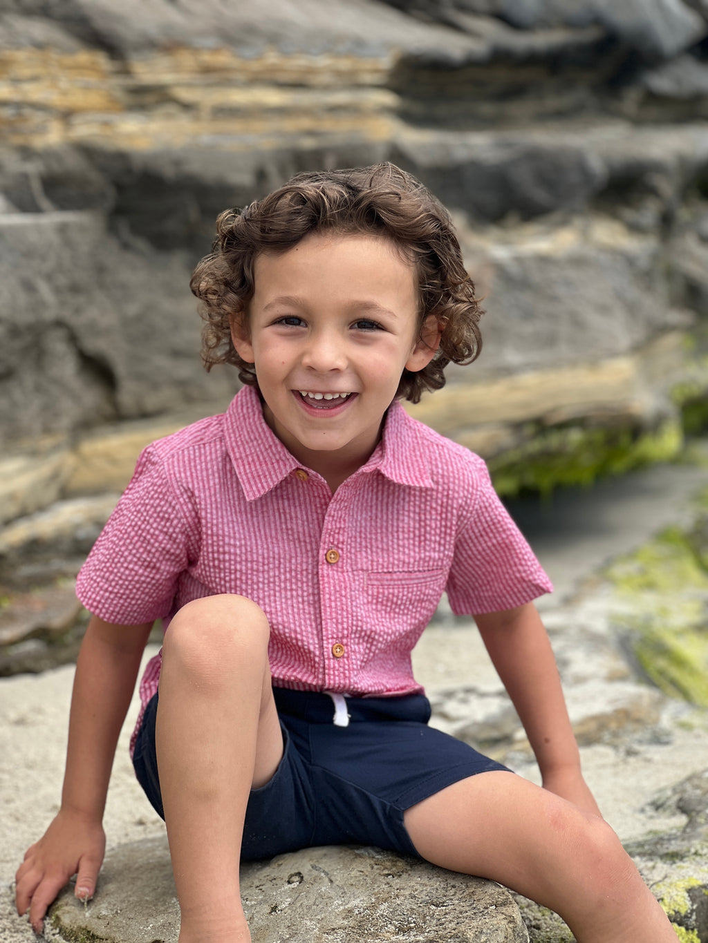 A boy with curly brown hair and brown eyes, wearing our Coral seersucker woven shirt and navy twill shorts sitting on a big rock at the beach