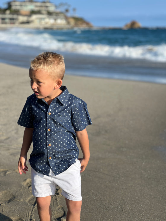 blonde hair boy wearing the chambray anchor woven shirt and white gauze shorts, walking in the sand at the beach in summer.