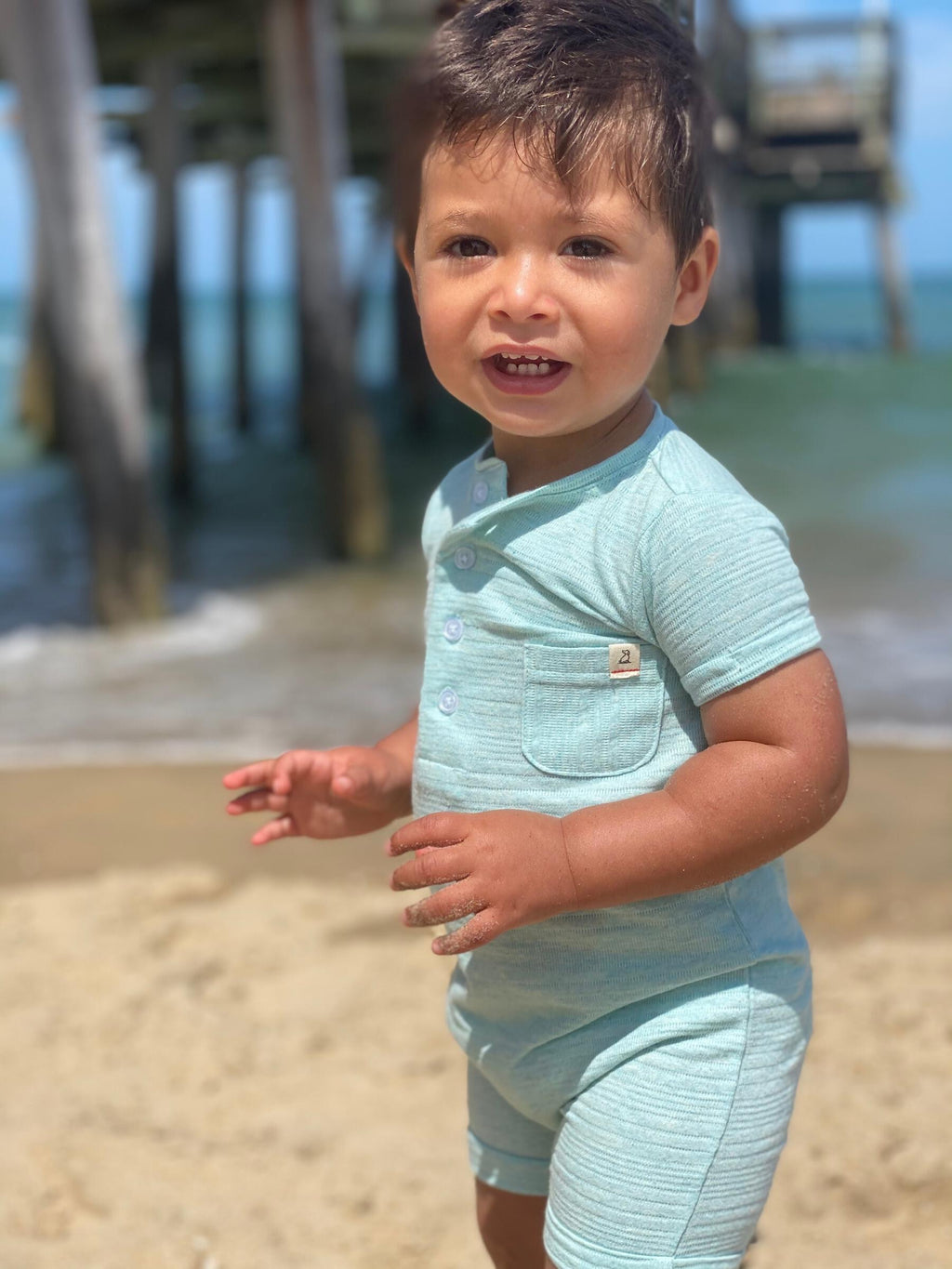 small boy withdrawn hair and brown eyes wearying the sky blue ribbed henley romper at the beach in summer.
