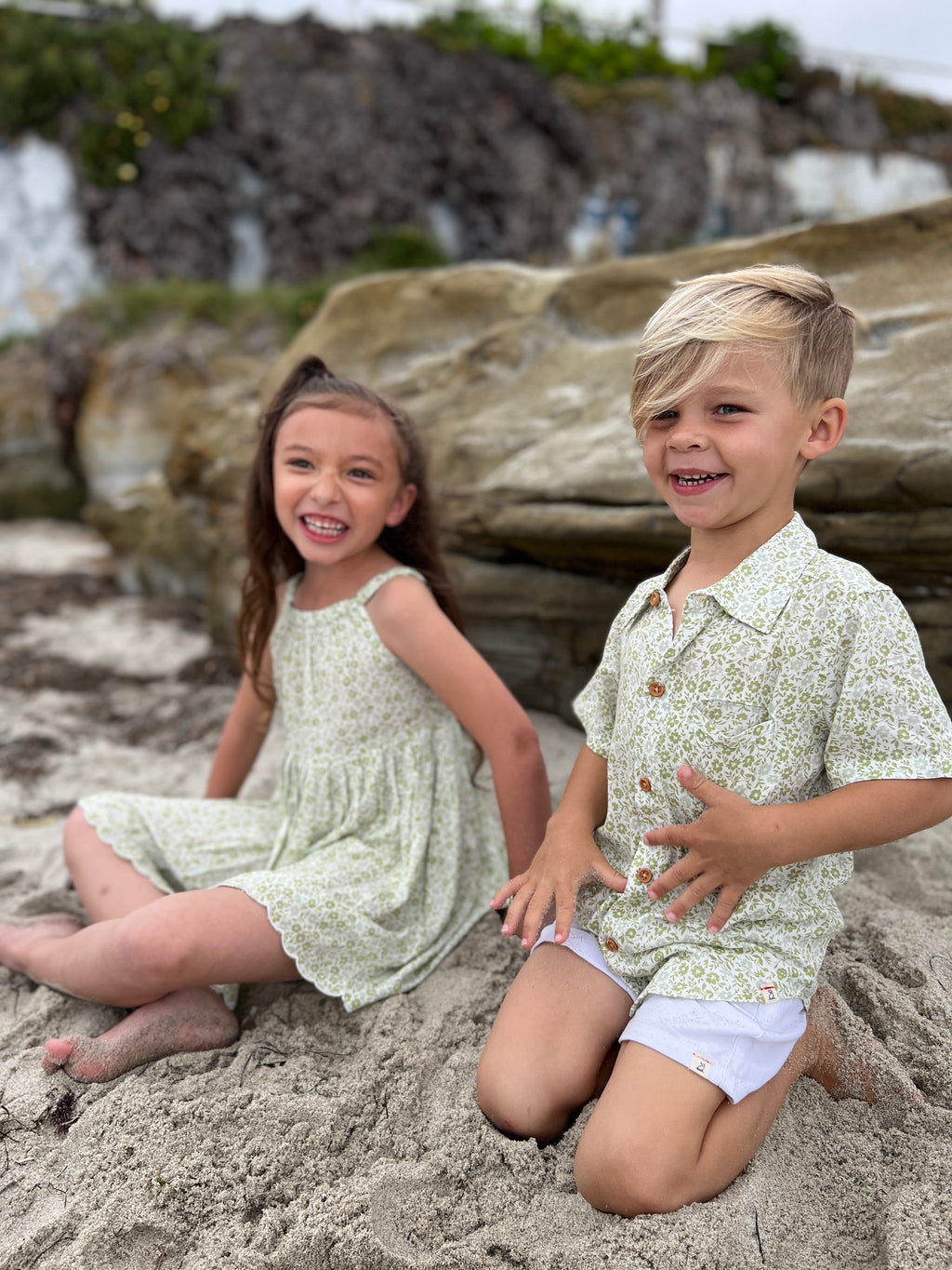 blonde boy with blue eyes wearing the green floral woven shirt and white gauze shorts, sitting on the sand with a girl with long brunette hair wearing a green floral vignette dress.