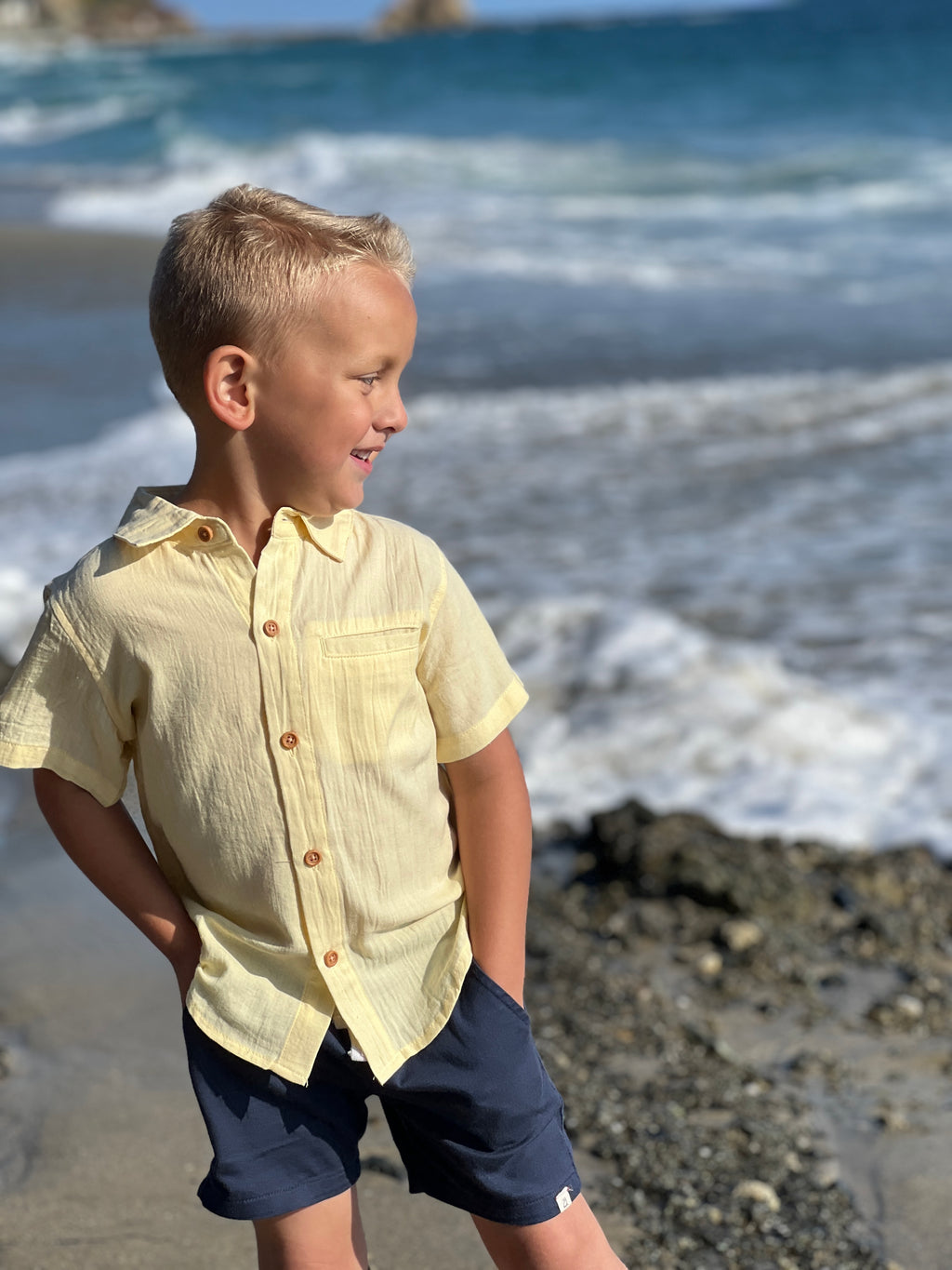 blonde hair boy wearing the lemon woven shirt and navy tiwill shorts by the sea at the beach.
