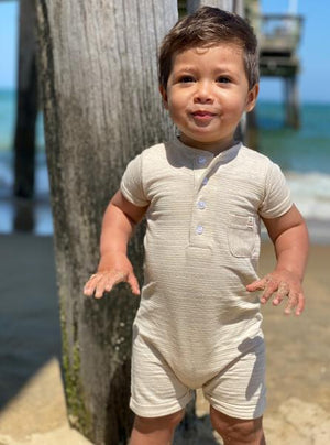 small boy with brown hair wearing beige ribbed henley romper, at the beach in summer