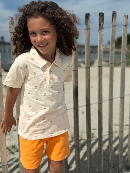 Curly haired boy in front of a fence wearing Short Sleeve Sleeved Sleeves Cream Henry Print Polo, Tangerine Terry Shorts.