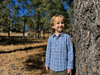 Young boy in a plaid shirt standing next to a tree in a forest
