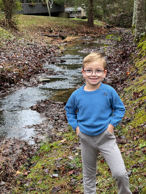 little blonde hair boy, circle glasses, woodland, small stream, autumn leaves, bright day, blue cotton sweater, knitted, 2 brown buttoms top left hsoulder, cuffed wrists, relaxed fit, layer, long sleeved, grey trousers