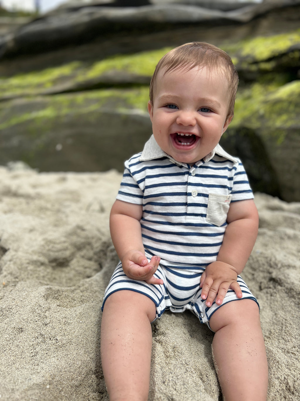Brown hair with blue eyes toddler wearing our navy/white polo romper sitting on the rocks at the beach .