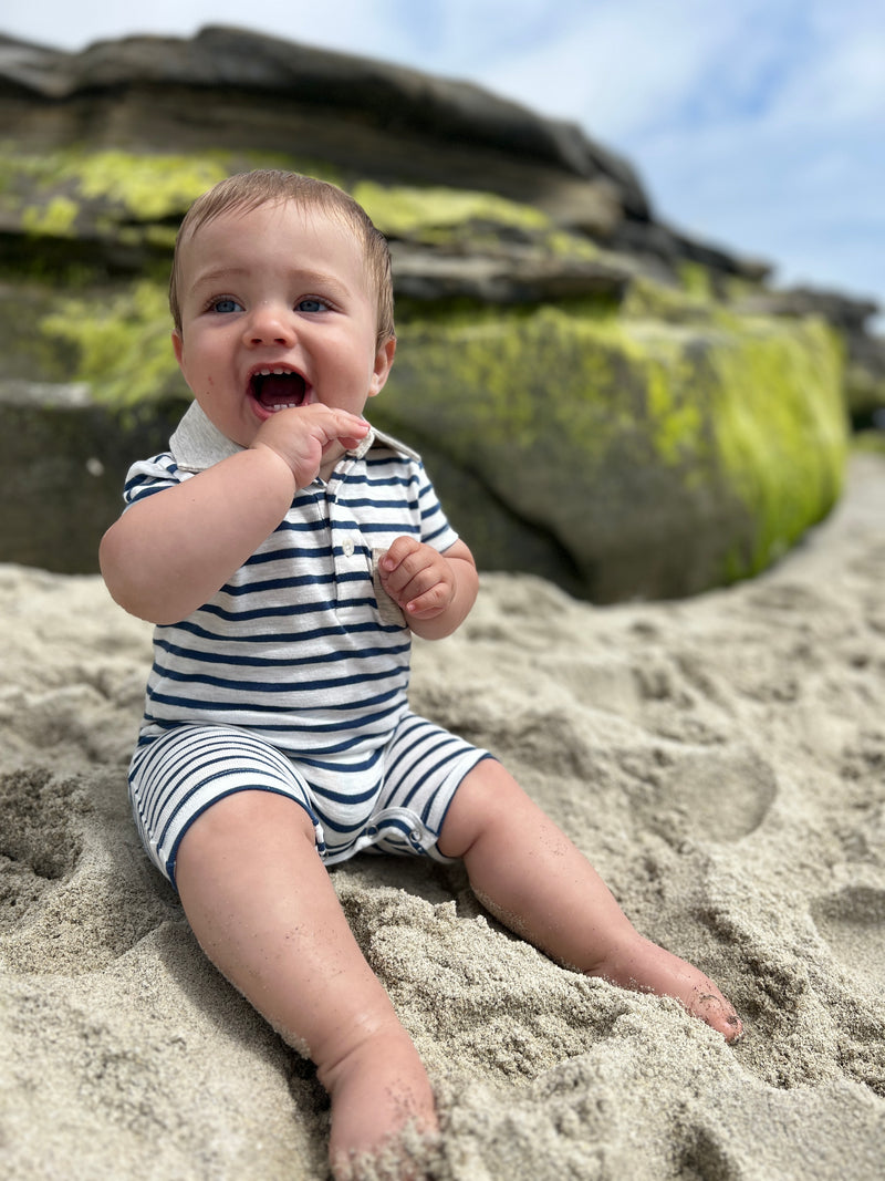 Brown hair with blue eyes toddler wearing our navy/white polo romper sitting on the rocks at the beach, smiling.