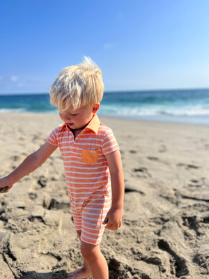 adorable blonde hair boy, wearing our peach/white polo romper , rolling a ball of sand in his hands at the beach in summer.