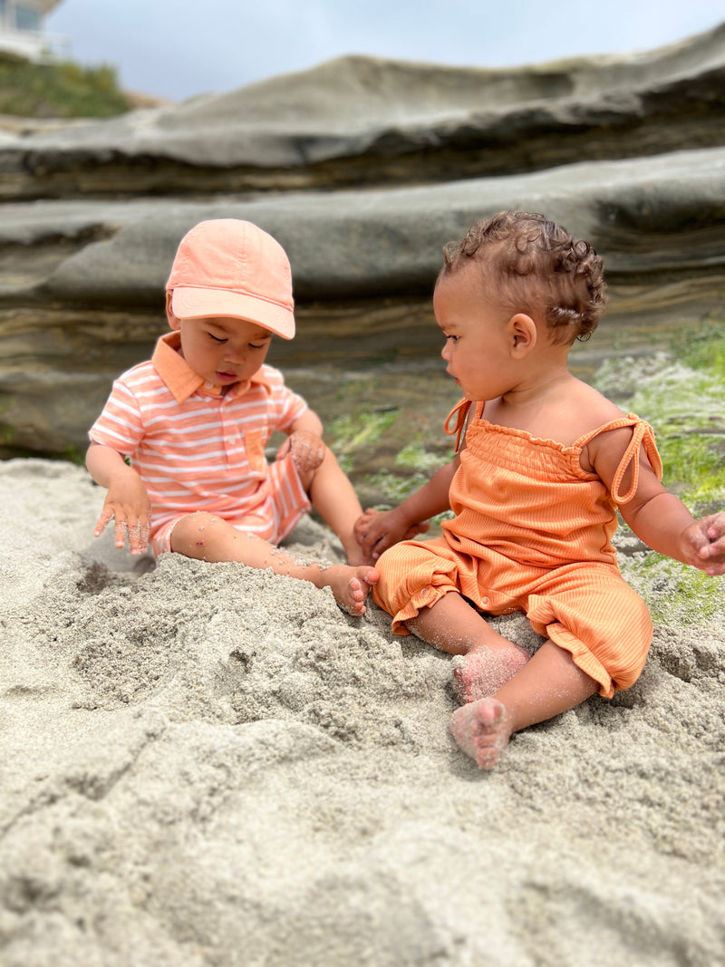 toddler wearing our peach/white polo romper and peach cap, playing the sand with a little girl with brown curly hair wearing a vignette orange frill playsuit