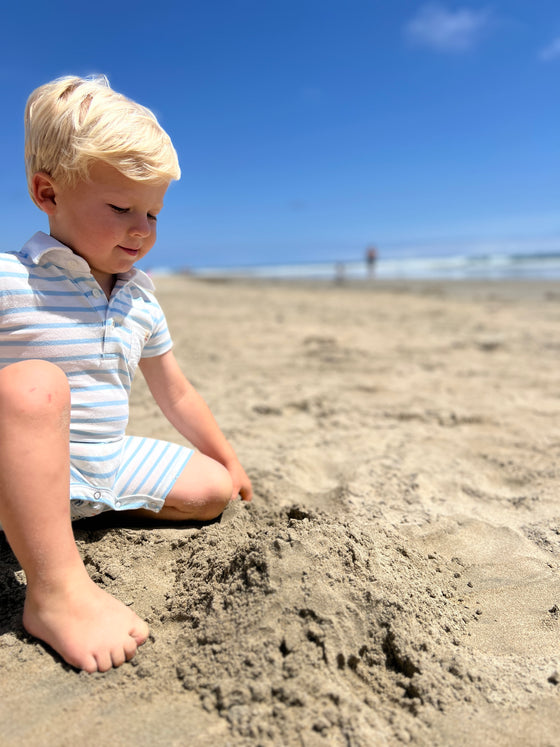 small blonde hair boy with eyes wearing our blue/white polo romper playing in the sand on the beach in the summer.