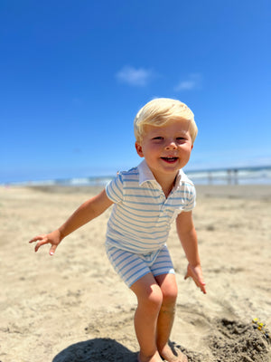 small blonde hair boy with eyes wearing our blue/white polo romper smiling while  playing in the sand on the beach in the summer.