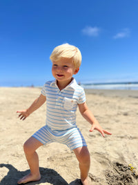 small blonde hair boy with eyes wearing our blue/white polo romper playing in the sand on the beach in the summer.