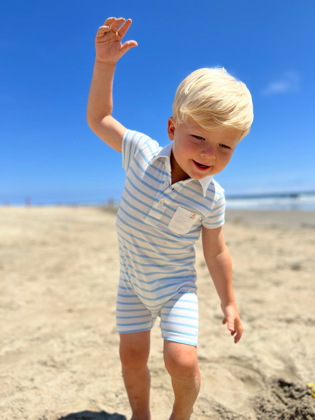 small blonde hair boy with eyes wearing our blue/white polo romper playing in the sand on the beach in the summer.