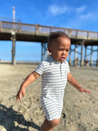 small boy with brown hair wearing our grey/white polo romper running along the beach in summer.