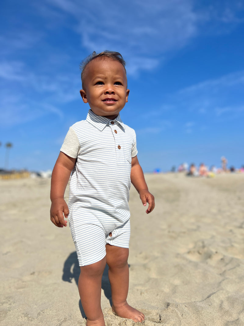 Toddler with brown hair and dark brown eyes wearing out Grey/white micro stripe romper standing on the sand at the beach insummer. 
