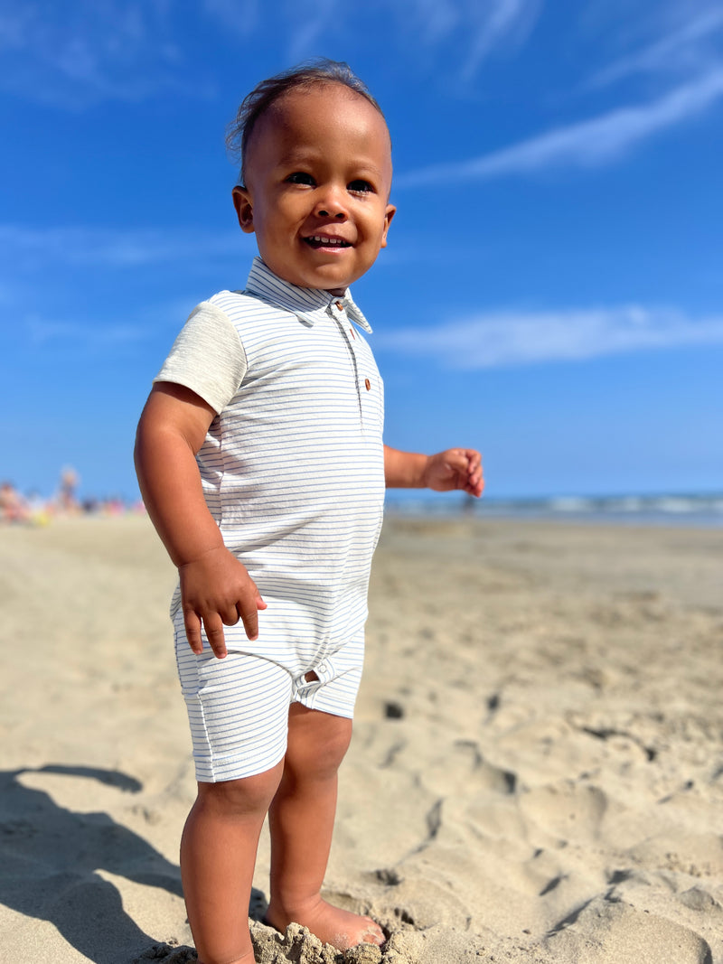 Toddler with brown hair and dark brown eyes wearing out Grey/white micro stripe romper standing on the sand at the beach insummer.