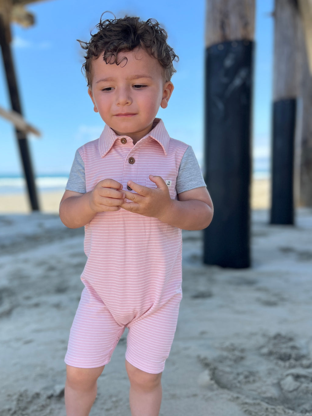 small boy with brown hair and brown eyes wearing our pink/white micro stripe polo romper standing under wooden bridge at the beach.