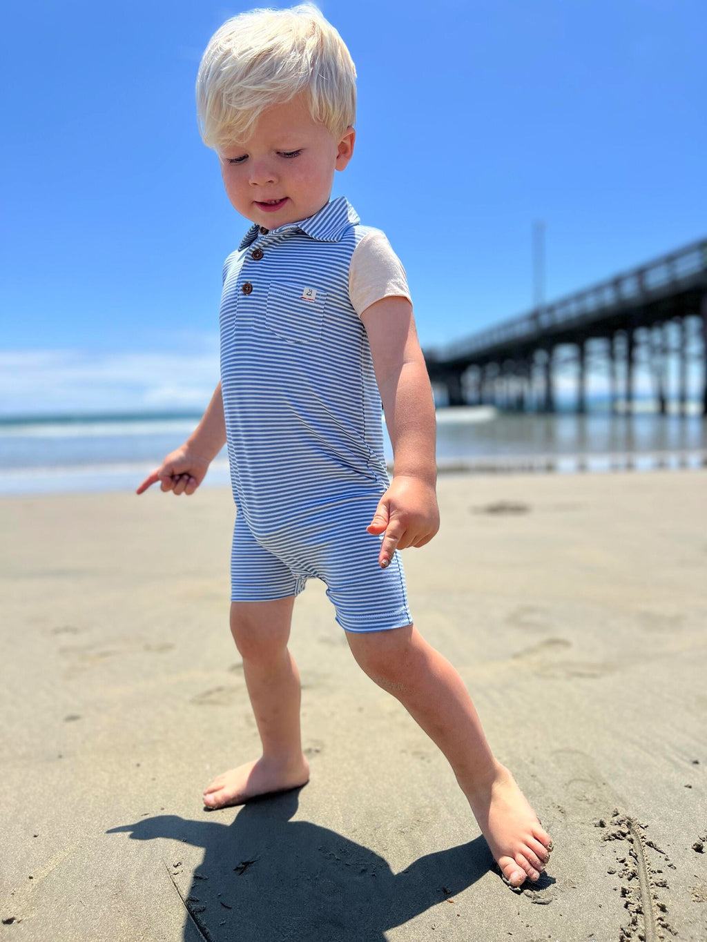 small blonde hair boy wearing our blue/white micro stripe polo romper, drawing in the sand with his feet at the beach