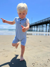 small blonde hair boy wearing our blue/white micro stripe polo romper, dancing in the sand at the beach in summer