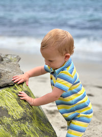little boy with blonde hair and blue eyes wearing the yellow/blue multi stripe polo romper standing on the sand next to a big rock on the beach