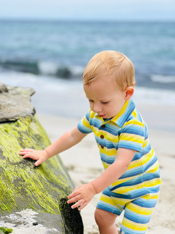 little boy with blonde hair and blue eyes wearing the yellow/blue multi stripe polo romper standing on the sand next to a big rock on the beach