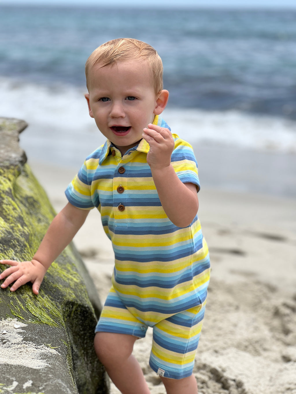 little boy with blonde hair and blue eyes wearing the yellow/blue multi stripe polo romper standing on the sand next to a big rock on the beach