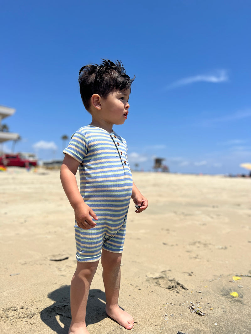 small boy with dark brown hair wearing our pink/white/grey ribbed henley romper standing on the beach looking out into the sea in summer.
