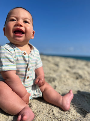 Adorable baby boy with brown hair and brown eyes, wearing our beige/lime/grey ribbed henley romper with a big smile, sitting on the sand at the beach in summer.
