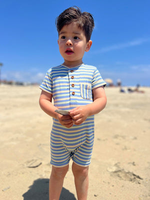 small boy with dark brown hair wearing our pink/white/grey ribbed henley romper standing on the beach looking out into the sea in summer.