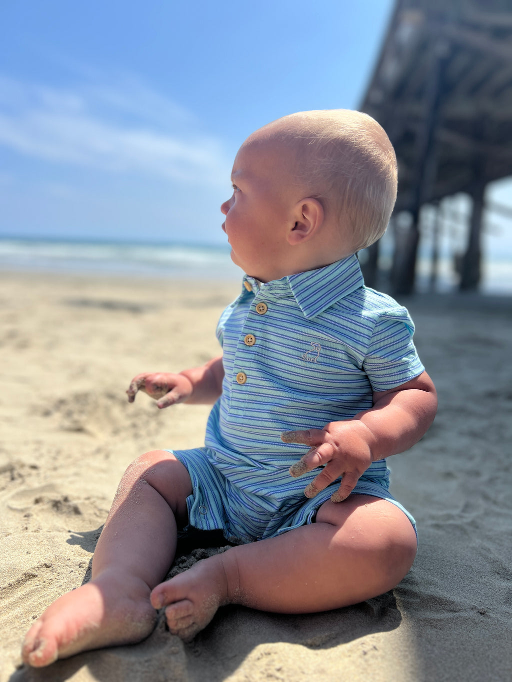 blonde hair baby boy sitting on sand at the beach wearing aqua blue stripe pique polo romper