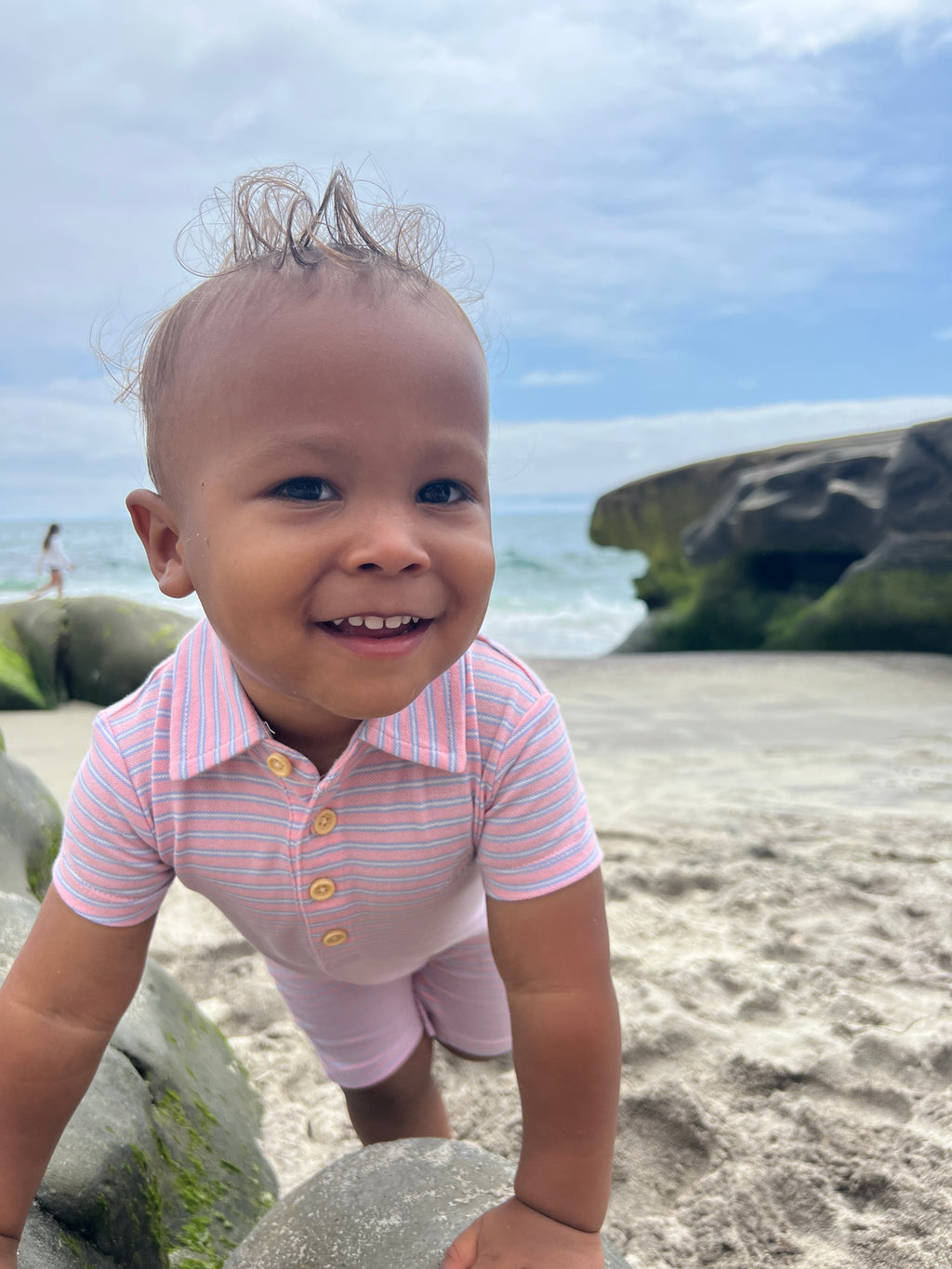 toddler with brown hair wearing our pink/lilac stripe pique polo romper, jumping onto the big rocks at the beach.