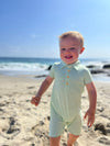 Small boy with blonde hair wearing our green/cream stripe pique polo romper , smiling while walking along the beach in summer.