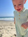 Small boy with blonde hair wearing our green/cream stripe pique polo romper , smiling 