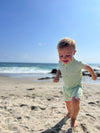 Small boy with blonde hair wearing our green/cream stripe pique polo romper , smiling while walking along the beach in summer.