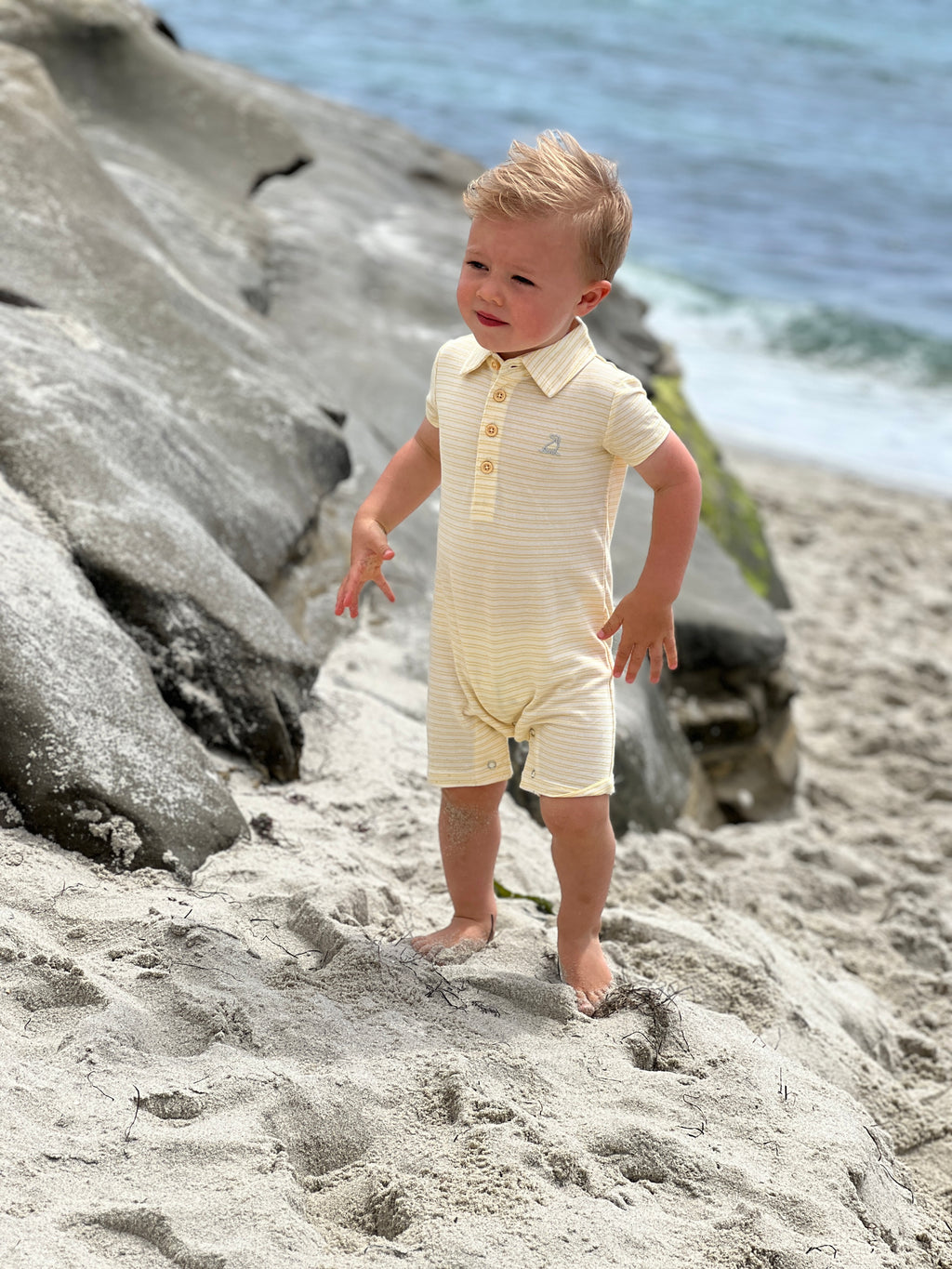 blonde hair, blue eyed boy wearing our yellow/cream stripe pique polo romper, standing in the sad at the beach in summer.