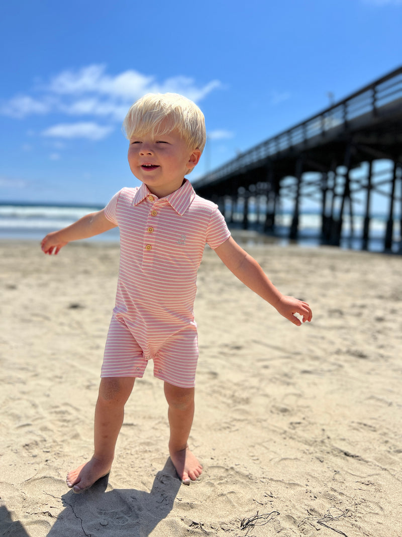 Small blonde hair boy wearing our cream/orange stripe pique polo romper standing in the sand at the beach in summer.