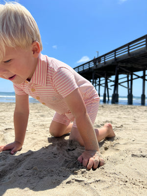 Small blonde hair boy wearing our cream/orange stripe pique polo romper playing with the sand at the beach in summer.