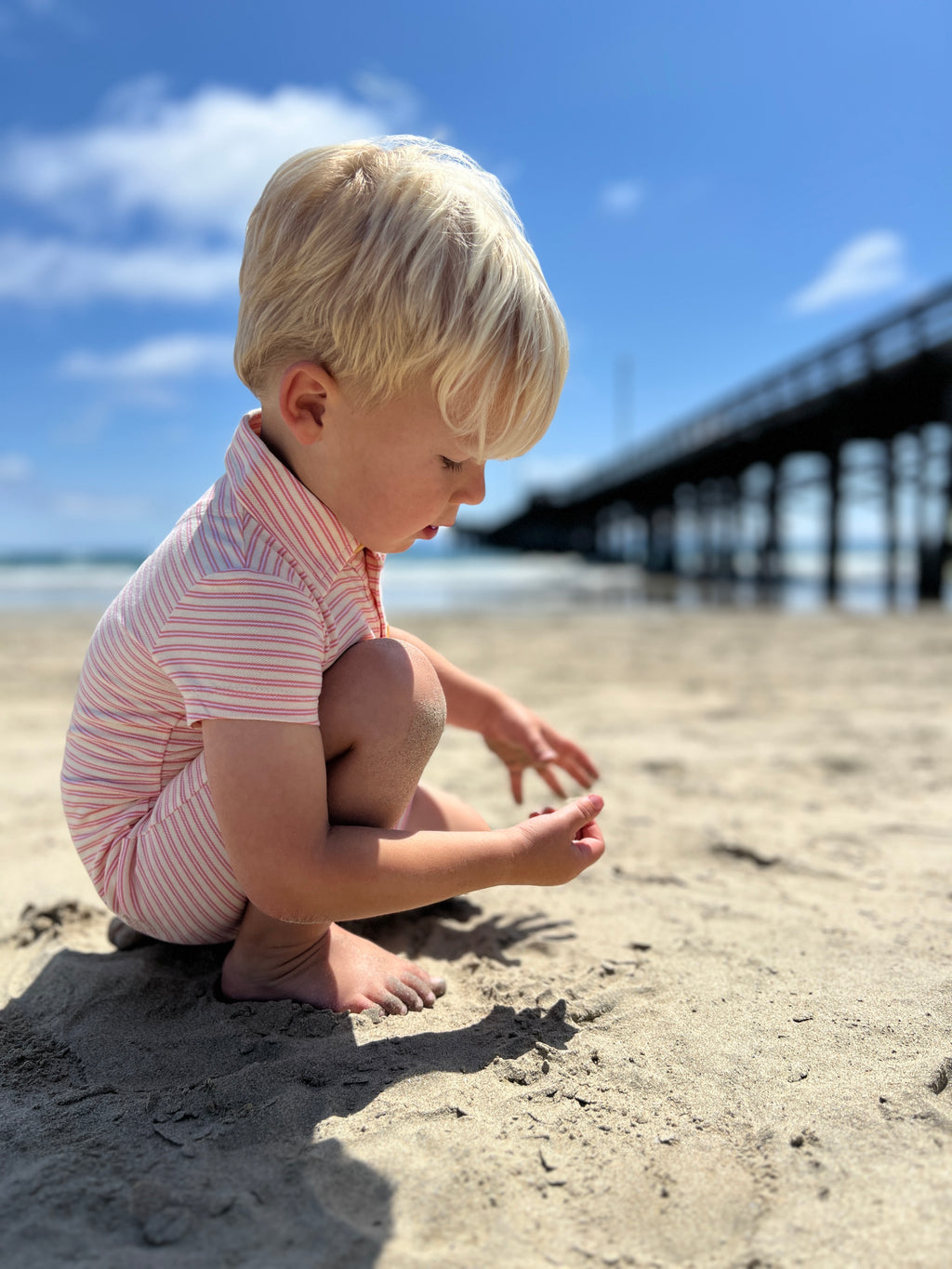 Small blonde hair boy wearing our cream/orange stripe pique polo romper playing with the sand at the beach in summer.